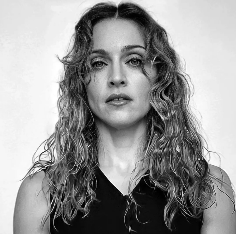 A black and white portrait of a woman with wavy, shoulder-length hair, looking directly at the camera with a serious expression. She wears a black sleeveless top and is set against a plain background.