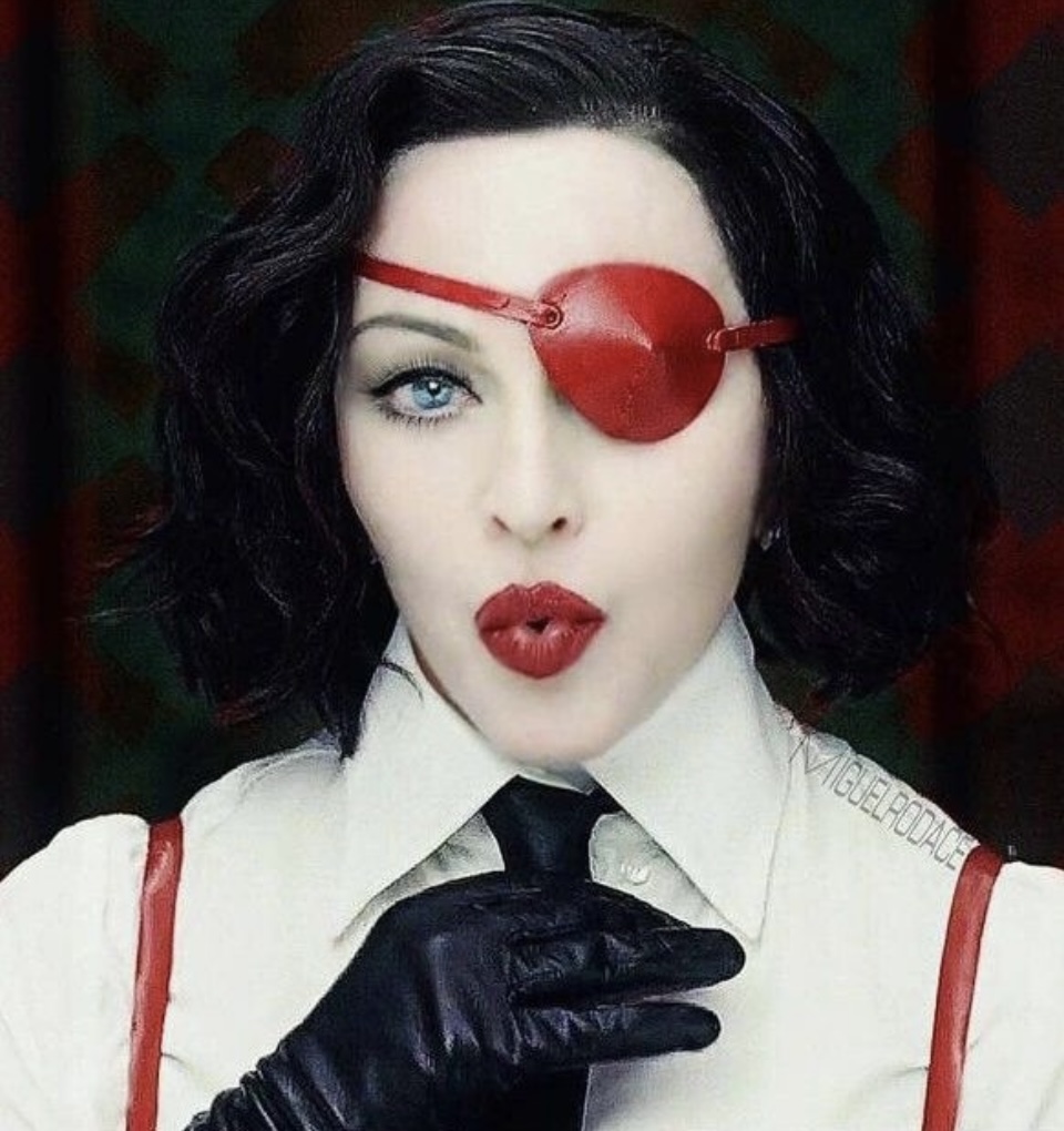 A woman with short black hair and bold red lips, wearing a white shirt and black gloves, poses with an exaggerated expression. She sports a red eye patch and has a striking gaze.