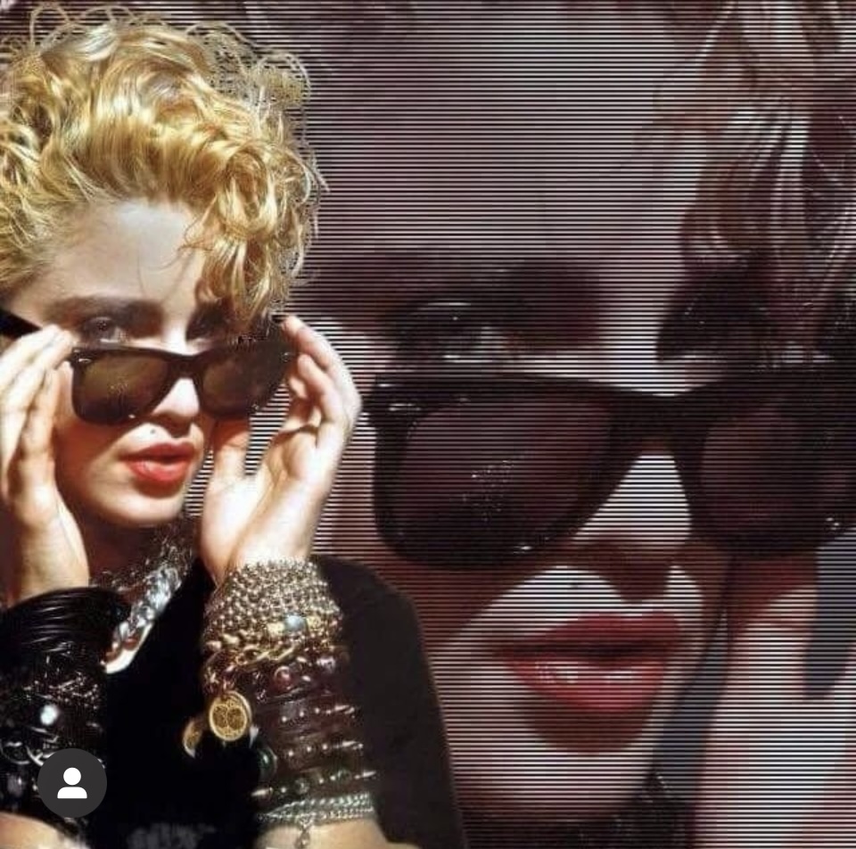 A woman with blonde curly hair wearing sunglasses, adorned with multiple bracelets, striking a confident pose. The background features a stylized, blurred image of another face with sunglasses.