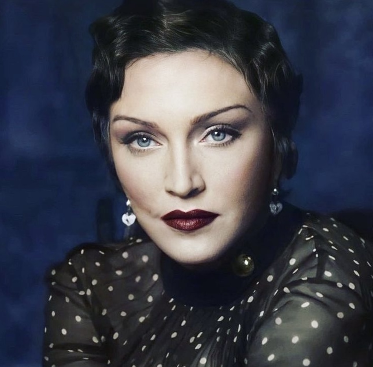 A close-up portrait of a woman with short dark hair, striking blue eyes, and bold red lips. She is wearing a sheer black blouse with white polka dots and heart-shaped earrings, against a dark blue background.