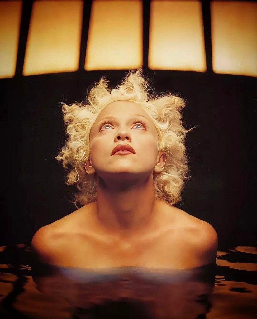A woman with curly blonde hair gazes upward while partially submerged in water, dramatic lighting highlighting her face and expression.