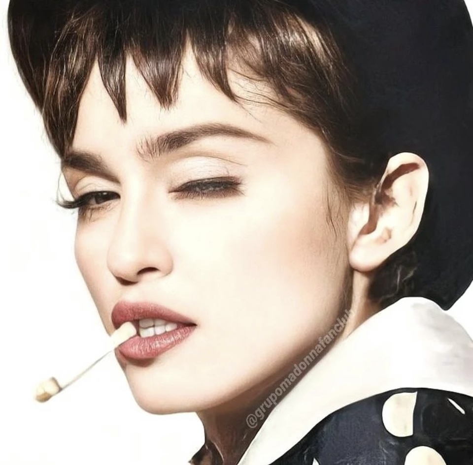Close-up of a woman with short hair, winking, and a toothpick in her mouth. She wears a black hat and a polka-dotted shirt.