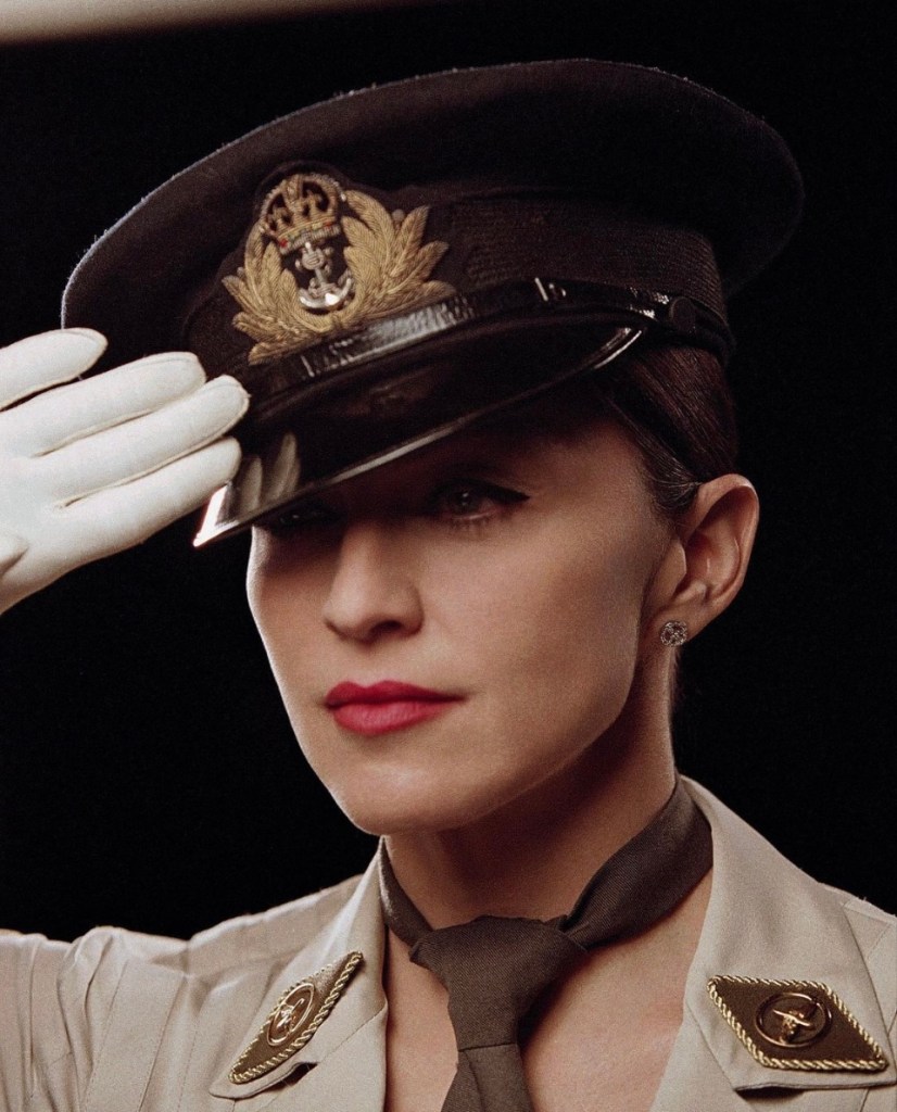 A woman in military attire saluting, wearing a peaked cap and white gloves. Her expression is serious, and she has bold red lipstick and distinct makeup.