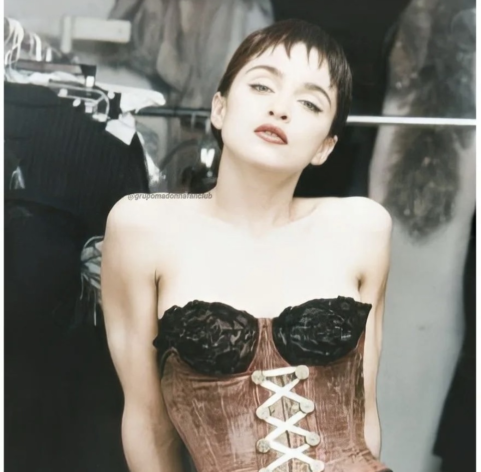 A young woman with short hair poses confidently in a corset with floral designs, standing in front of a clothing rack.