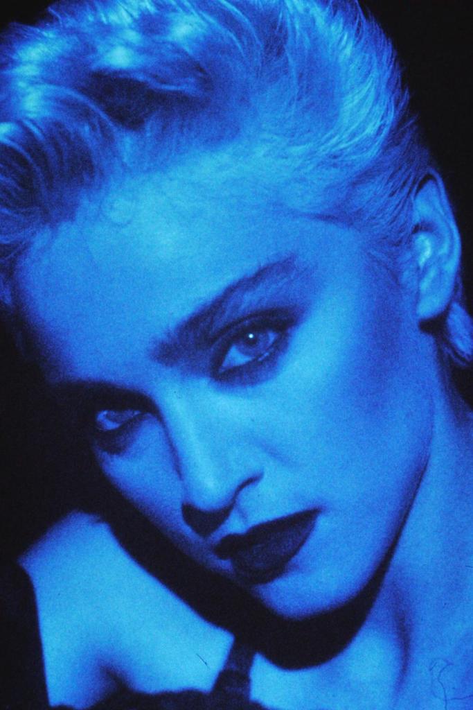 Close-up portrait of a person with platinum blonde hair, dramatic blue lighting, and bold makeup, featuring dark lipstick and striking eyes.