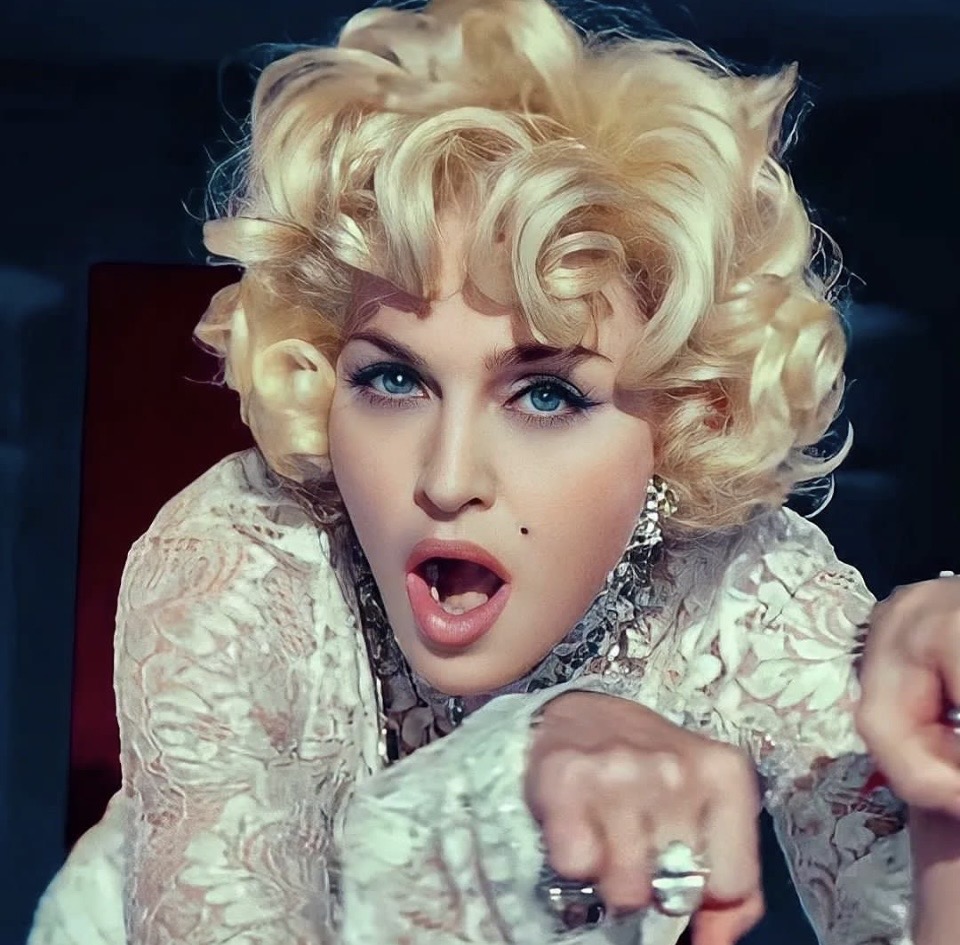 A close-up image of a woman with blonde, curly hair and striking blue eyes, wearing a white lace outfit and jewelry, expressing excitement or surprise.