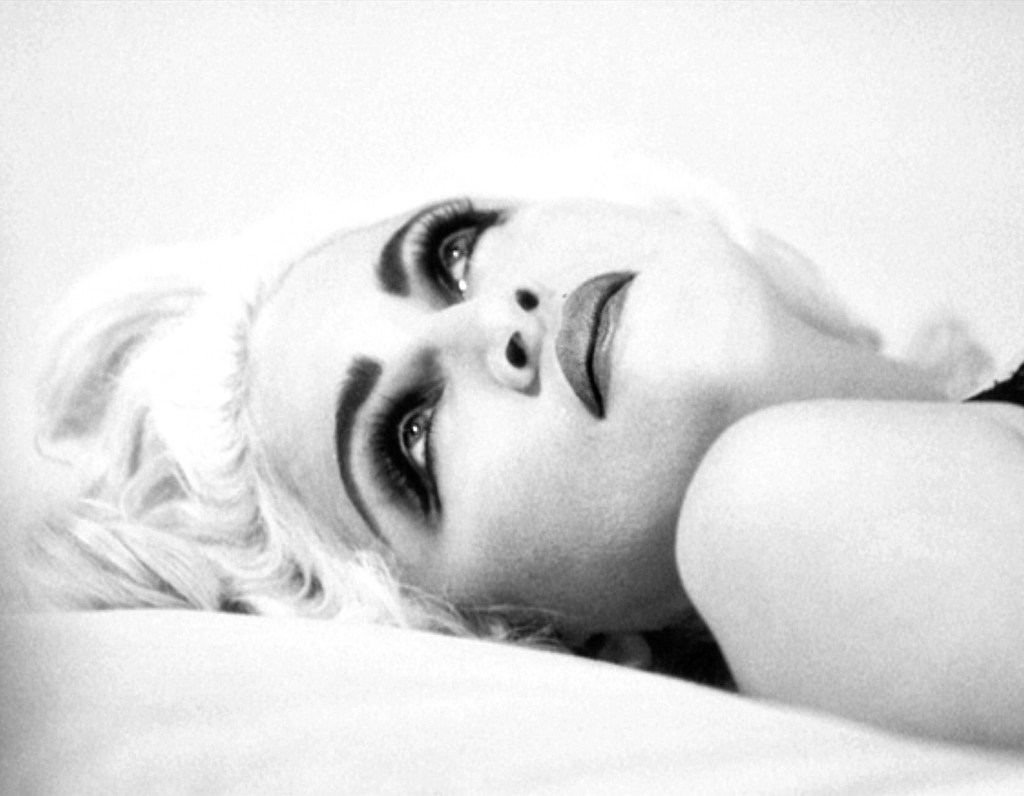 A black and white portrait of a woman lying down, gazing upward with striking makeup and blonde hair.