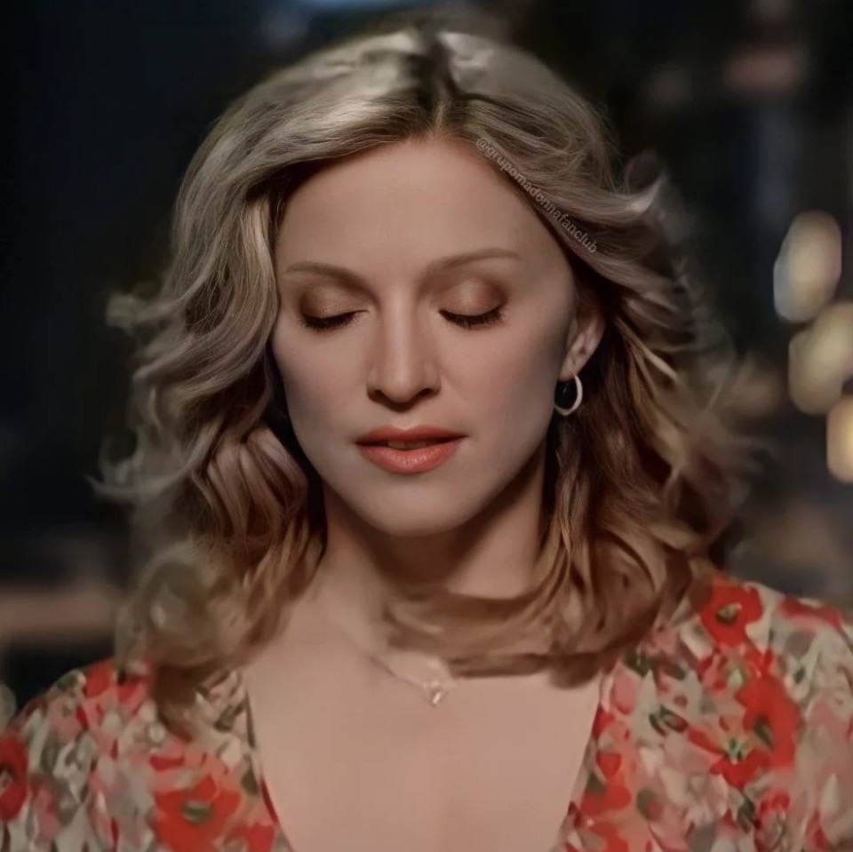 A woman with wavy, blonde hair and a floral dress has her eyes closed, with a serene expression on her face.
