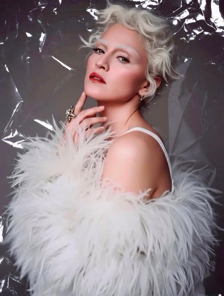 A woman with short, curly platinum hair poses in profile, wearing a white feathered coat. She has a subtle makeup look and is touching her face with one hand, set against a shiny, crinkled backdrop.