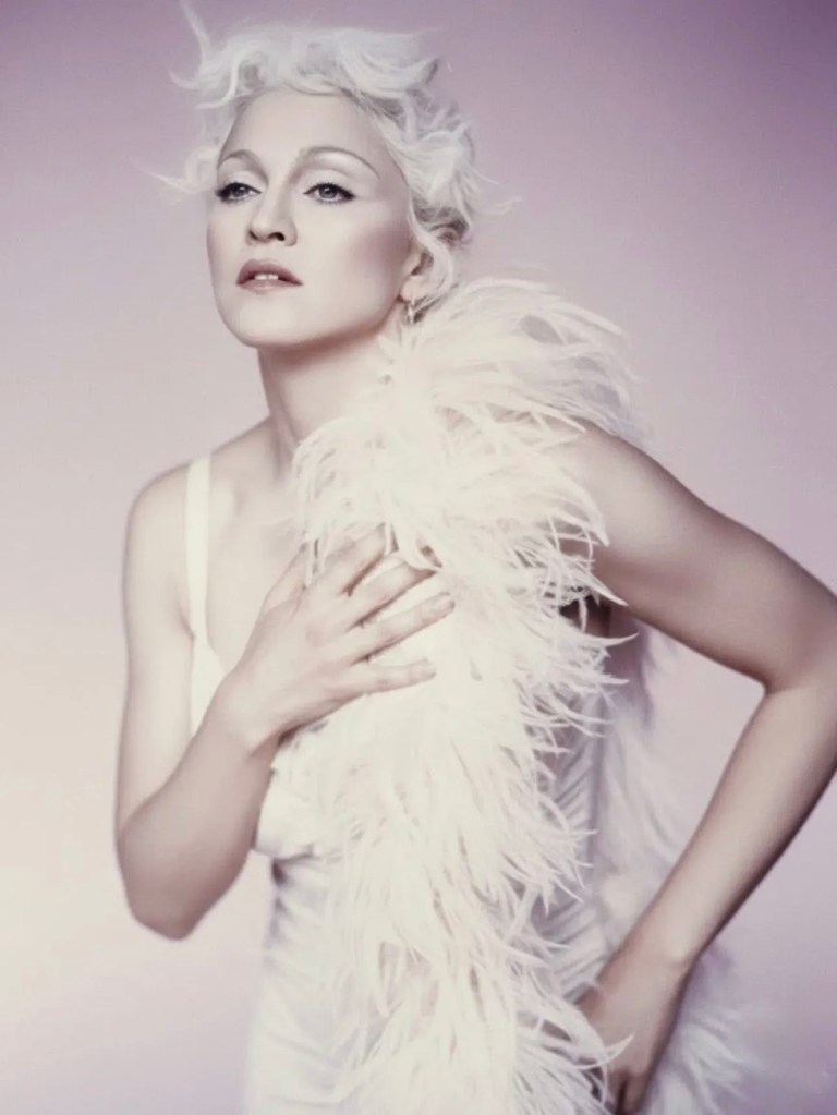 A woman with styled short white hair wearing a white dress and a white feather boa, posing against a soft pink background.