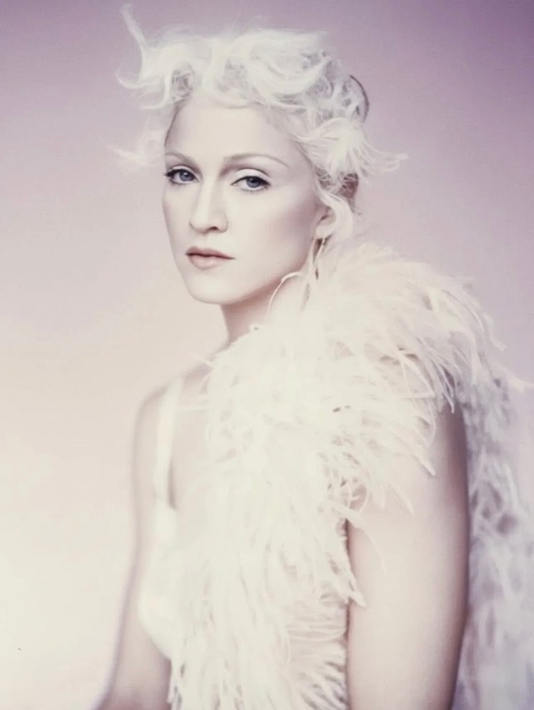 A close-up portrait of a woman with striking white hair styled in soft curls, wearing a feathered white garment and a subtle makeup look, against a pale background.