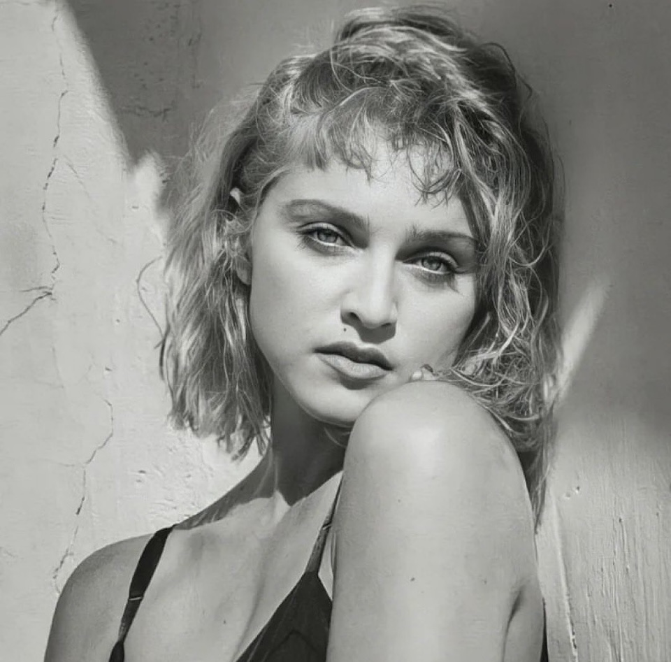 Black and white portrait of a young woman with short, wavy hair, looking directly at the camera with a serious expression against a textured wall.