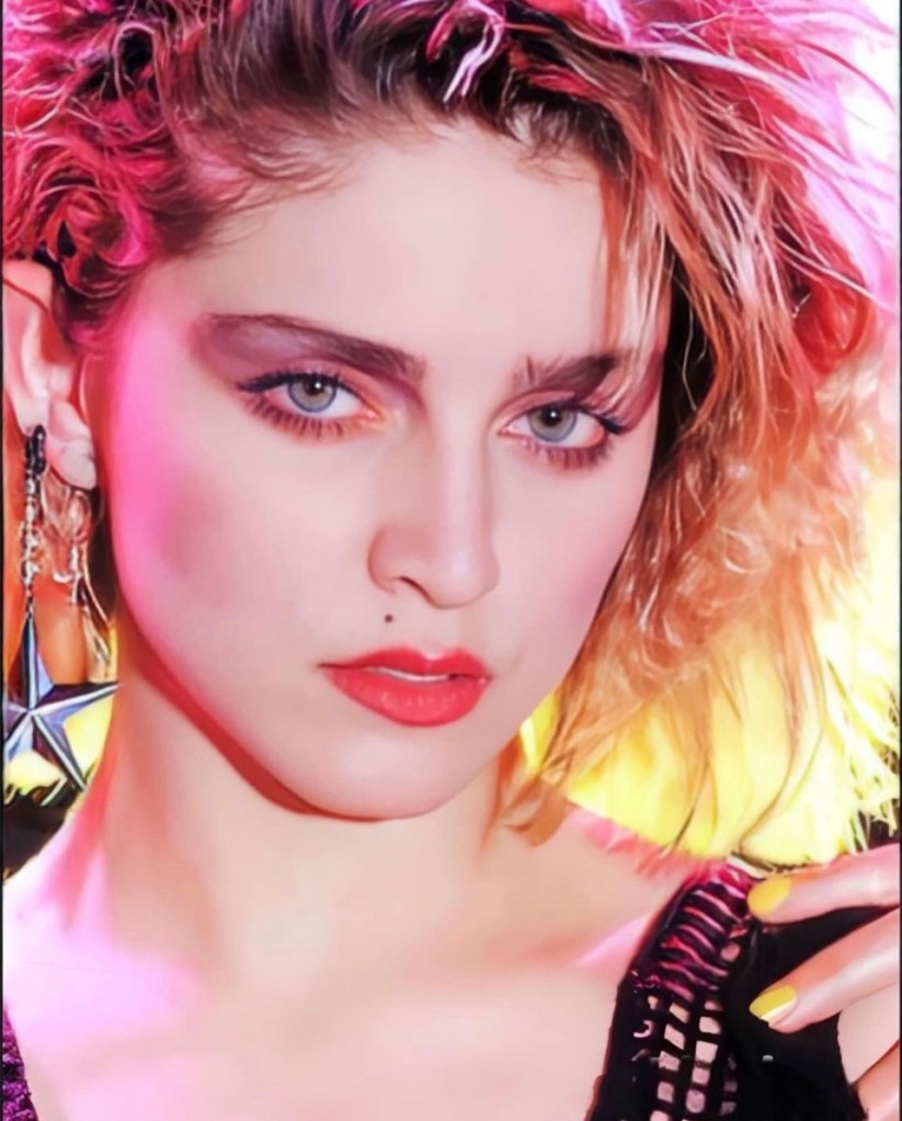 Close-up portrait of a woman with curly blonde hair, bright blue eyes, and bold makeup, featuring pink and orange hues. She wears statement earrings and has a playful expression, with one hand on her shoulder.