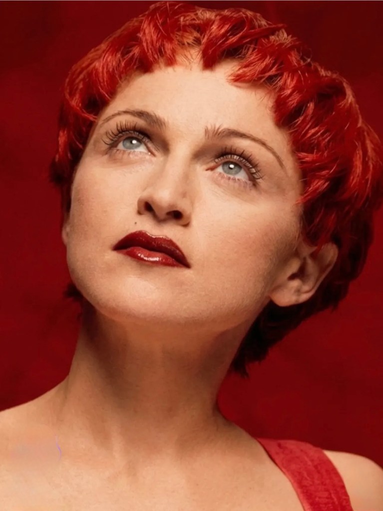A woman with short red hair looking up with a pensive expression against a red background.