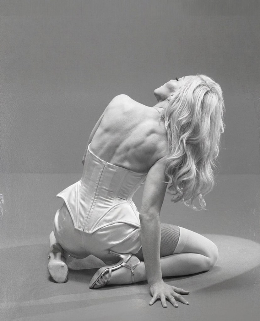 A woman in a corset and ballet shoes poses elegantly on her hands and knees, showcasing her back and long hair in a monochrome setting.