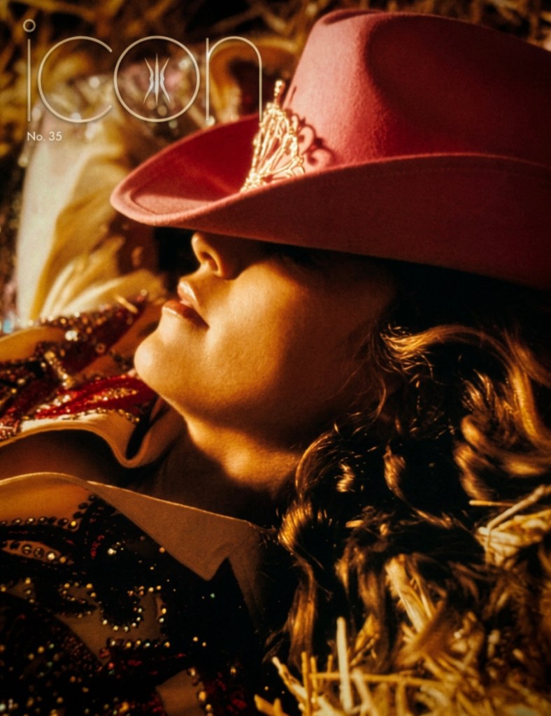 A close-up of a woman lying on straw, wearing a pink cowboy hat adorned with a tiara. She has long, wavy hair and is dressed in a sequined top with intricate designs.