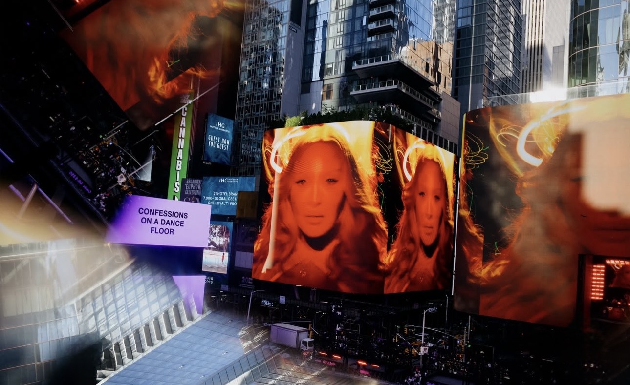A vibrant display in Times Square featuring large LED screens showcasing a close-up of a woman with long hair, illuminated with a warm orange hue, alongside a sign that reads 'Confessions on a Dance Floor'. The background includes tall buildings and bustling street activity.