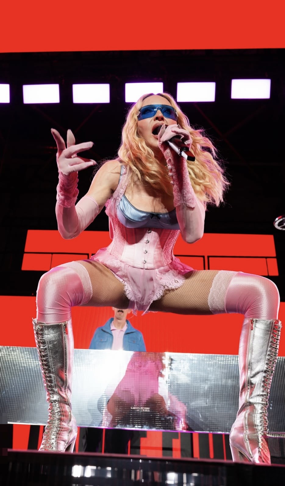 A performer in a provocative pink outfit and silver knee-high boots sings passionately on stage, with vibrant red lighting in the background.