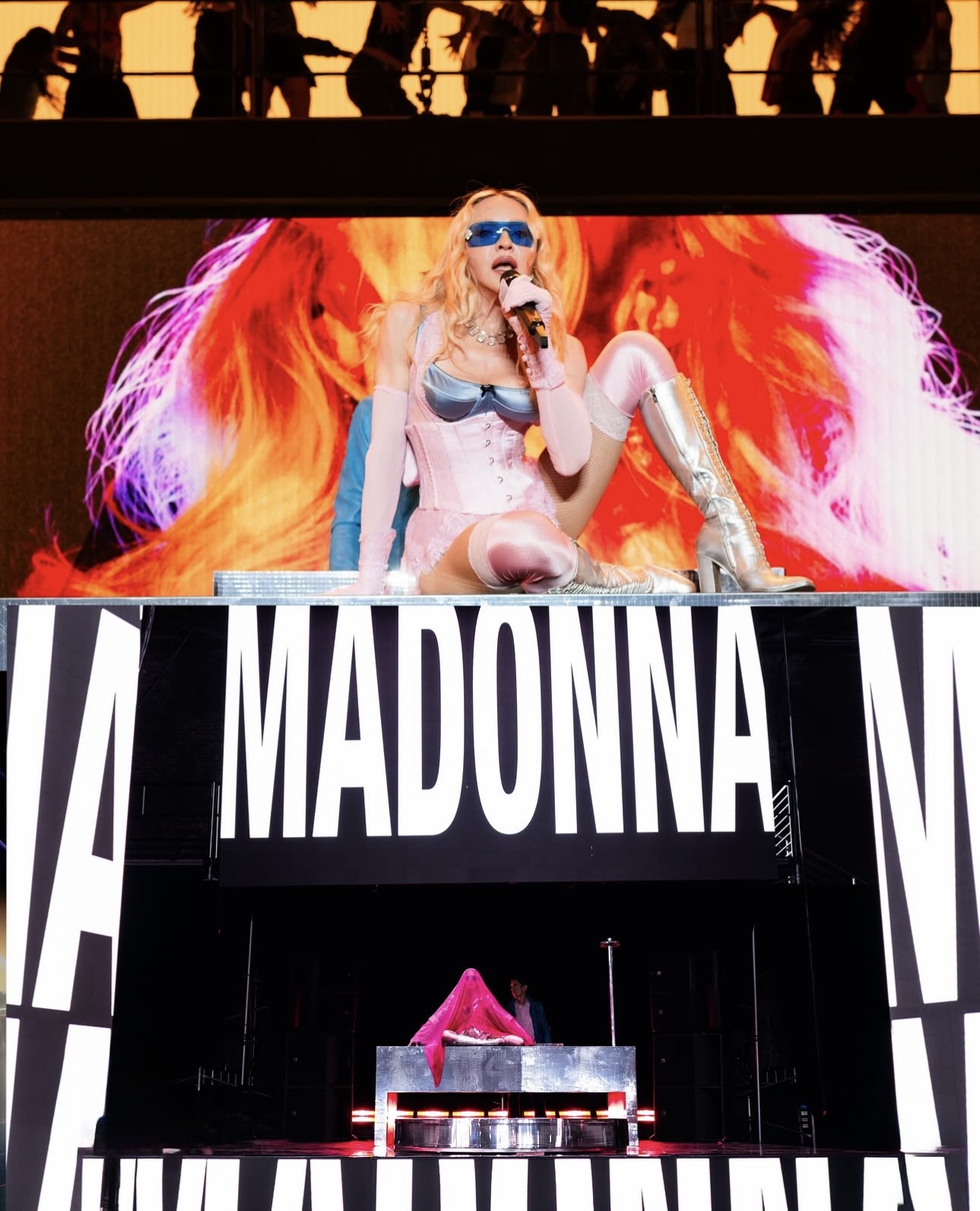 A performer with long blonde hair, wearing a pink outfit and sunglasses, sings into a microphone on stage with a vibrant digital backdrop displaying pink and purple graphics.