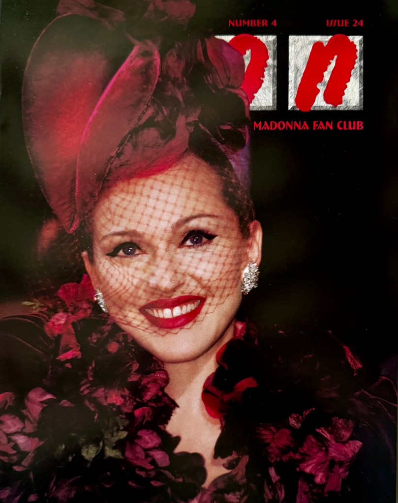 A smiling woman wearing a large hat adorned with flowers and a black veil, surrounded by colorful floral decorations, featured on the cover of a Madonna fan club magazine.