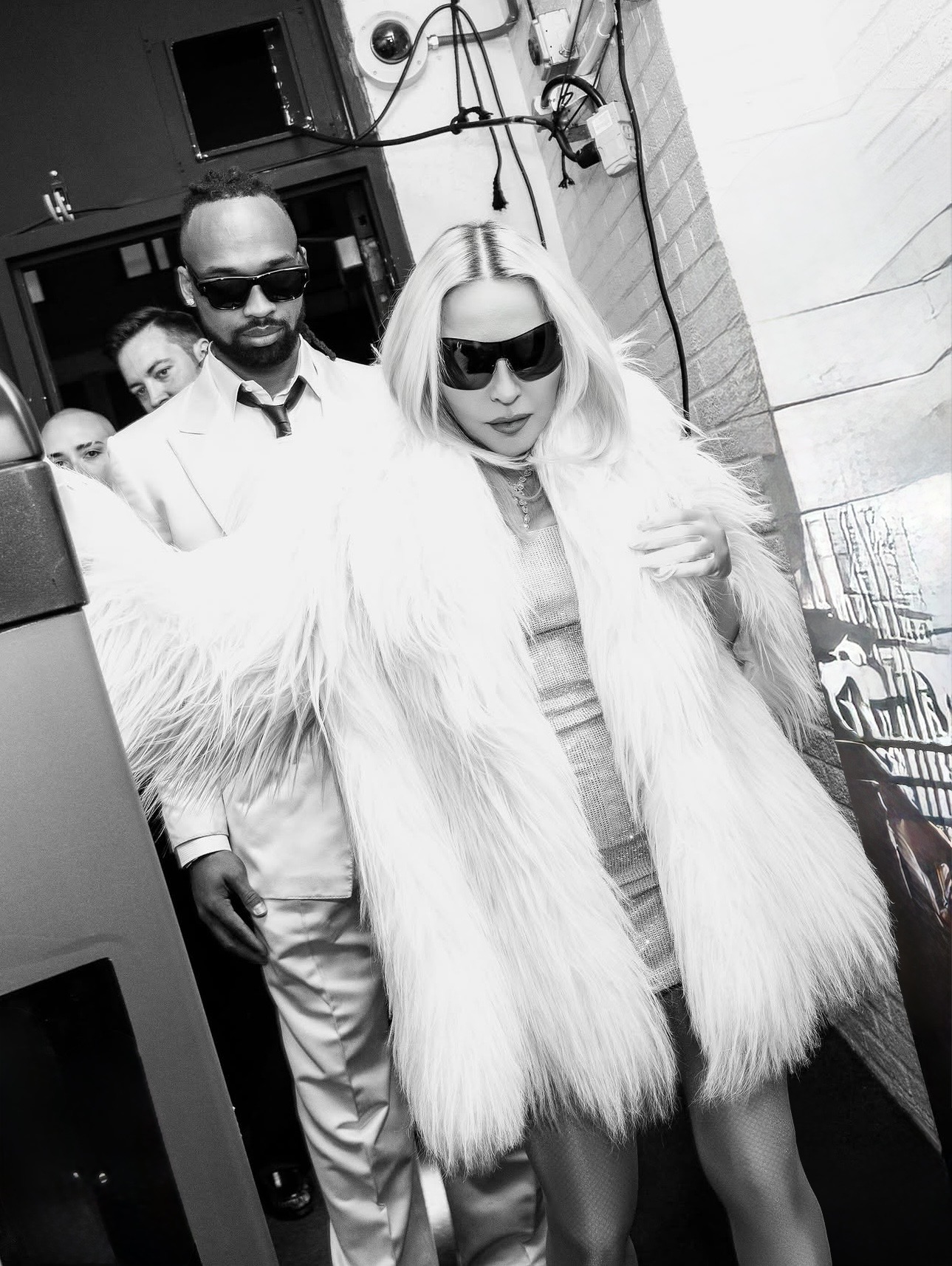 A stylish woman in a white fur coat and sunglasses walks confidently, accompanied by a man in a white suit and sunglasses, against a backdrop of an urban environment, creating a glamorous and mysterious atmosphere.