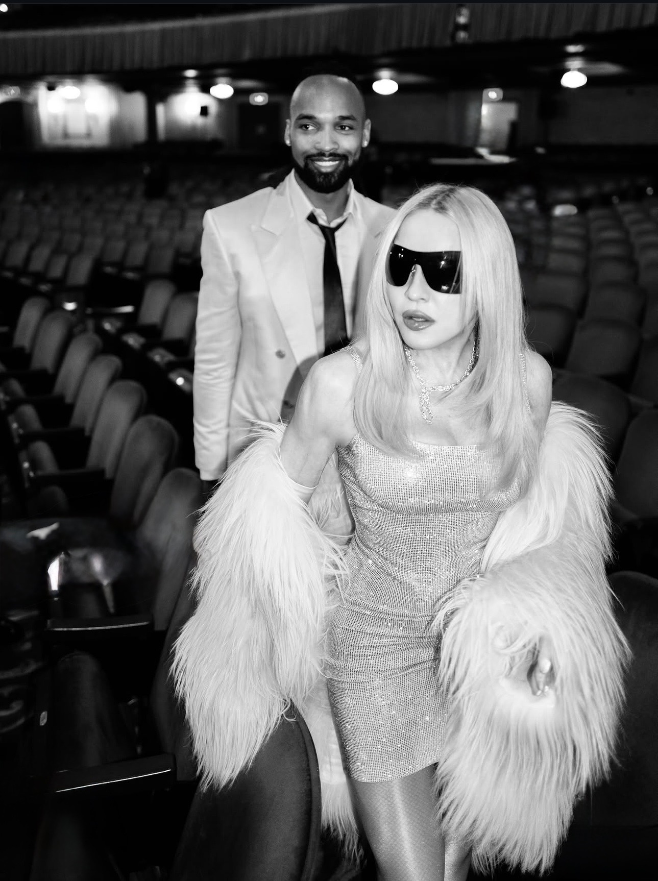 A fashionable couple in a theater, with a man in a light suit and a woman in a sparkly dress wearing sunglasses and a fur coat. The setting is dimly lit with empty seats in the background.