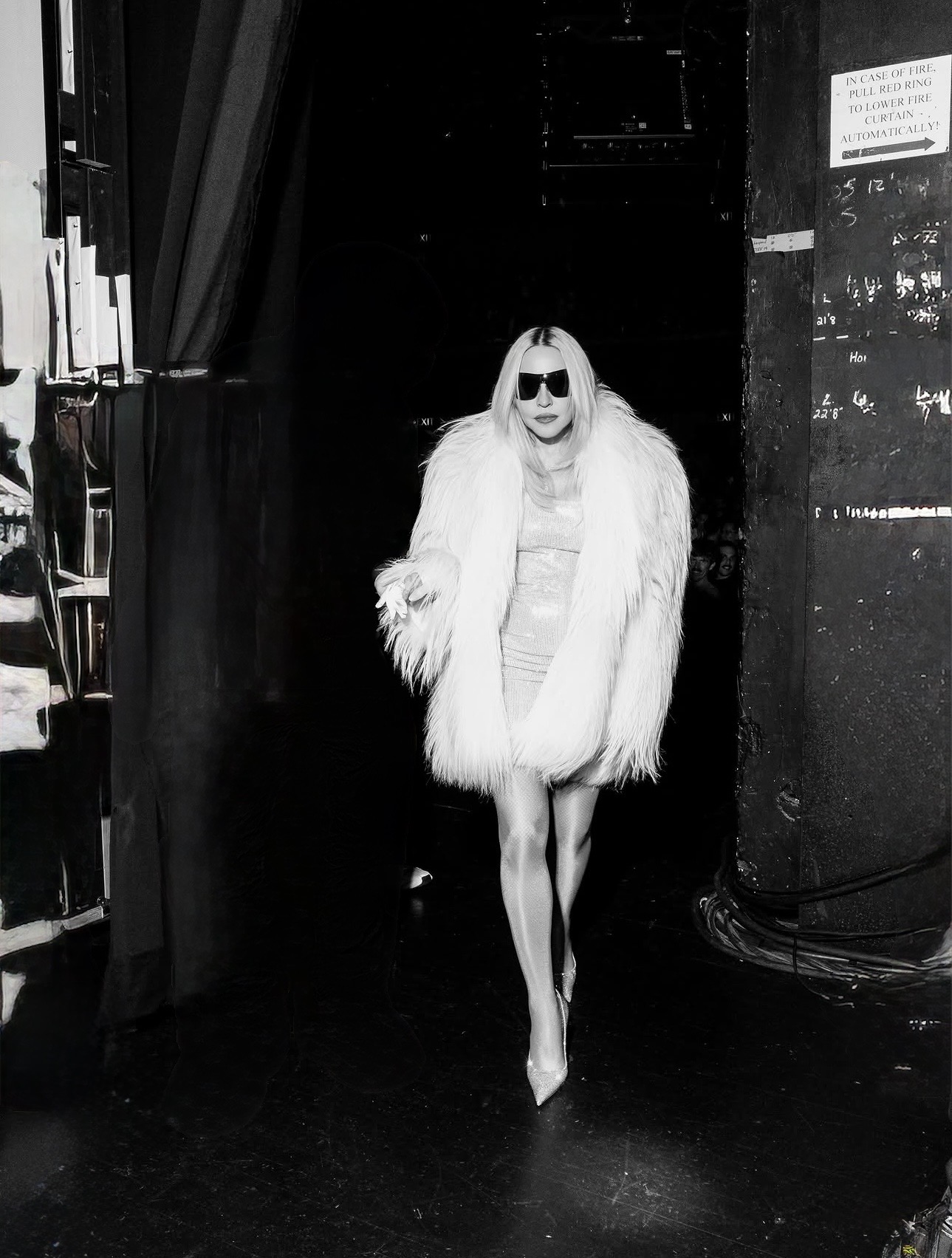 A glamorous figure in a white fur coat and sunglasses walks onto a stage, with a dramatic backdrop of black curtains and stage lighting.