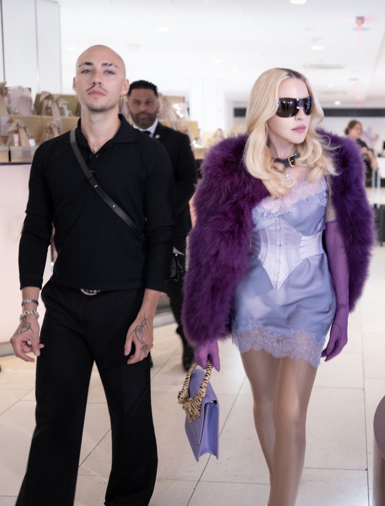 A stylish man in a black outfit with tattoos walking alongside a woman in a glamorous outfit featuring a purple fur coat, tinted sunglasses, and a lavender dress, holding a small handbag, as they navigate a retail space.