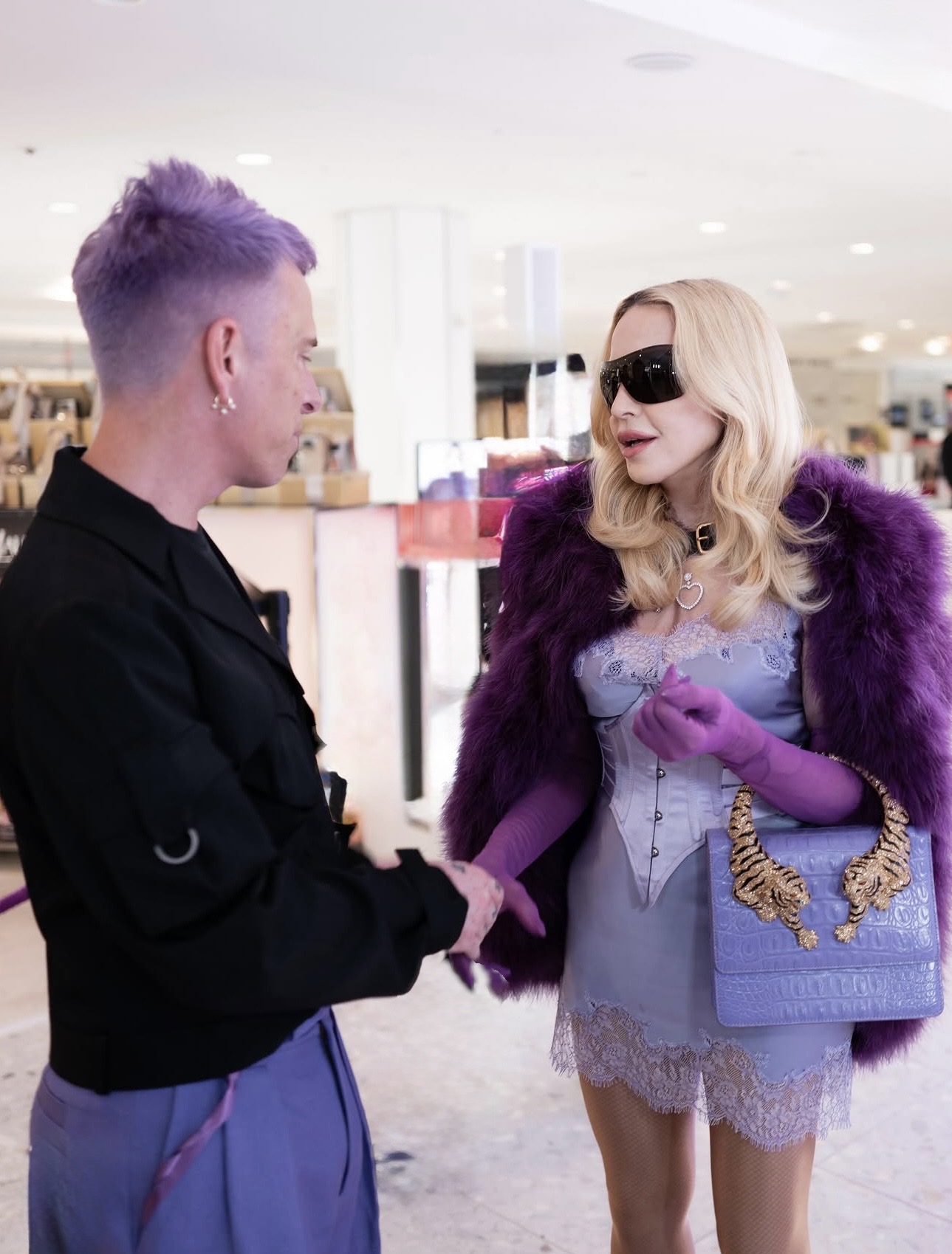 Two stylish individuals engage in conversation in a retail setting. One person has purple hair and wears a black outfit, while the other has long blonde hair, sunglasses, and a glamorous purple coat.