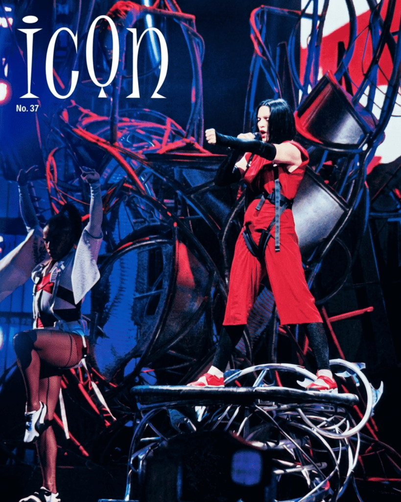 A performer in a striking red outfit sings on stage, surrounded by dynamic lighting and intricate set design, while a second performer dances nearby.