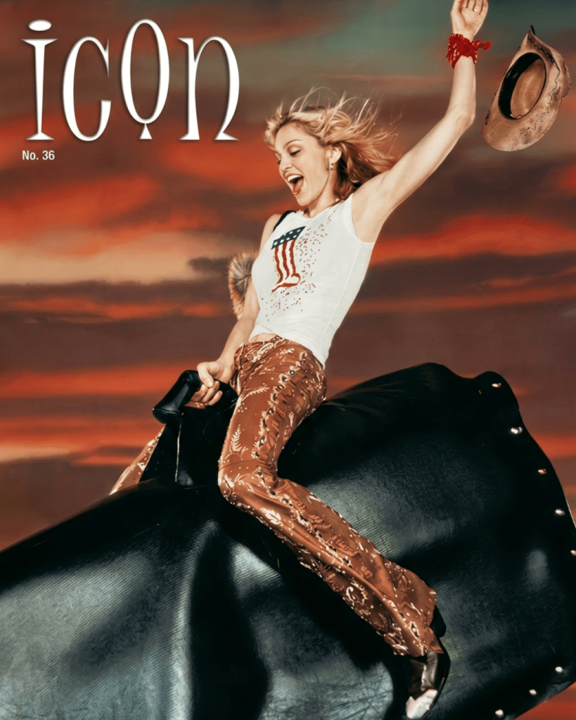 A woman with long hair is joyfully riding a mechanical bull, wearing a white tank top with an American flag design and brown patterned pants. She is raising one hand in excitement, and a cowboy hat is flying in the air.