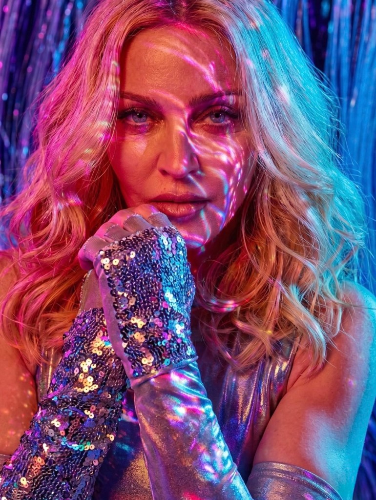 Close-up of a woman with wavy blonde hair, wearing a shiny silver outfit and sequined gloves. Colorful light patterns are cast on her face, creating an artistic and vibrant effect.