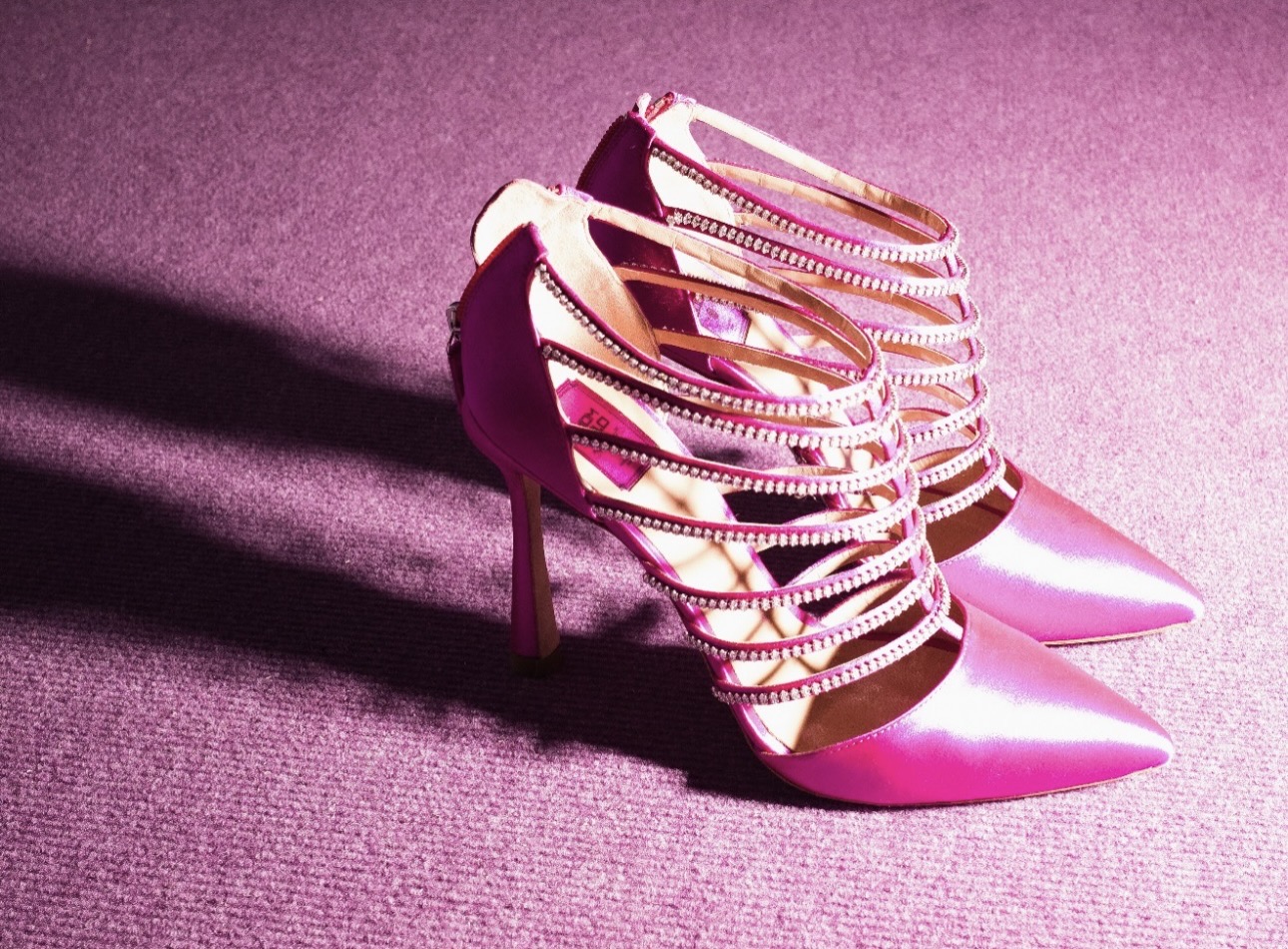 A pair of stylish pink high-heeled shoes with crisscross straps and metallic accents, placed on a purple surface.