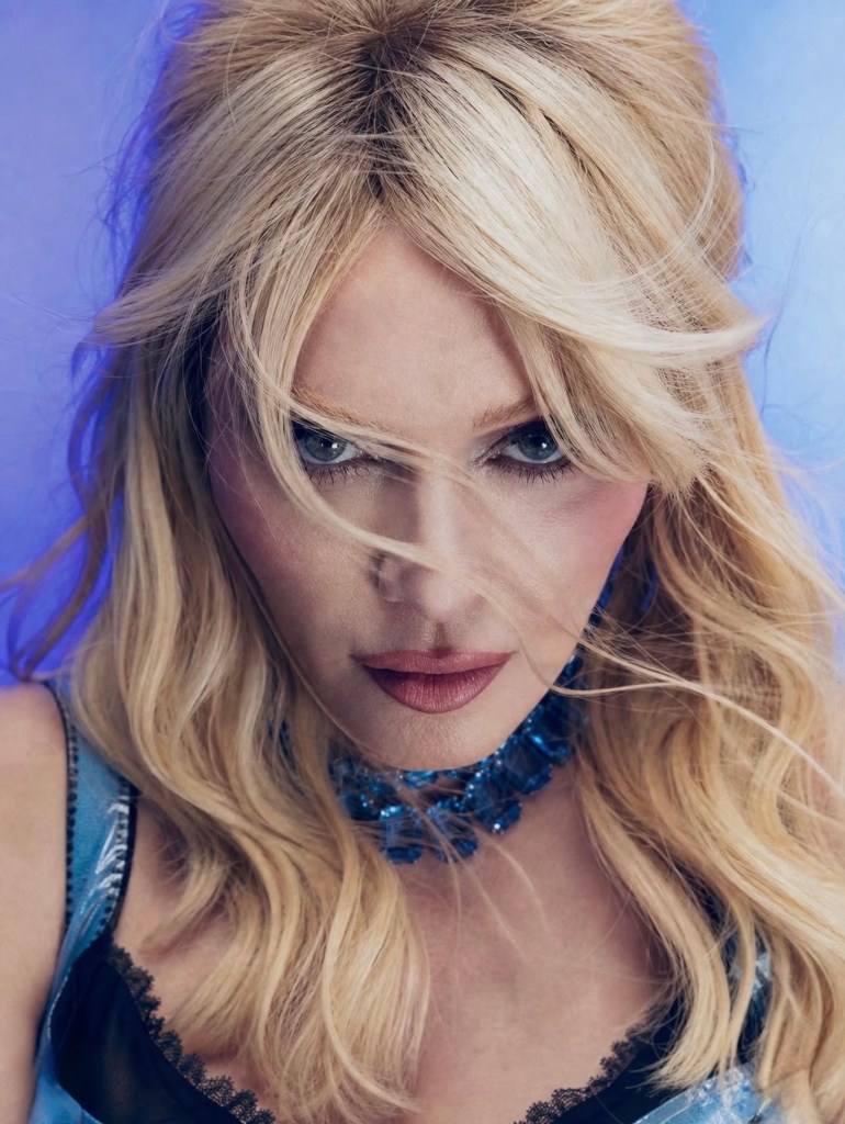Close-up portrait of a person with long, wavy blonde hair and a blue statement necklace, looking directly at the camera against a soft blue background.