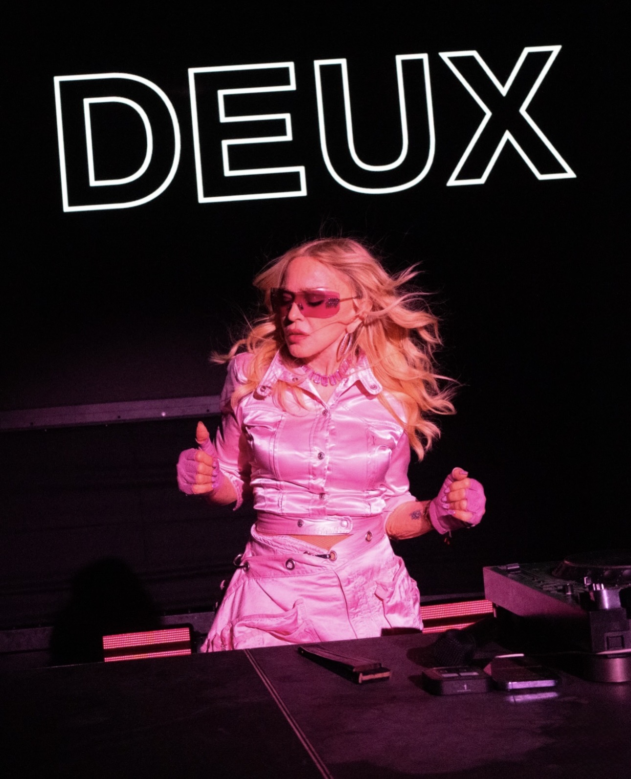 A performer in a pink outfit with sunglasses, energetically engaging with an audience, against a dark background featuring the word 'DEUX'.