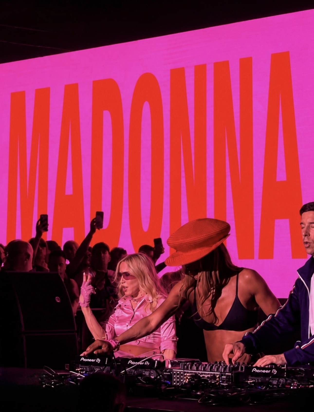 A vibrant stage scene featuring three DJs at turntables, with a large pink backdrop displaying 'MADONNA'.